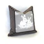 Cushion Two is better, square cushion in gray velvet with an illustration of two white rabbits in the center, framed by a black ribbon and black buttons at the corners.