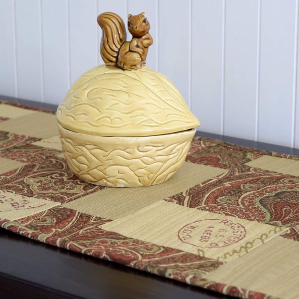 Détail du chemin de table À l'aventure déposé sur un meuble avec une noix et un écureuil en céramique.
