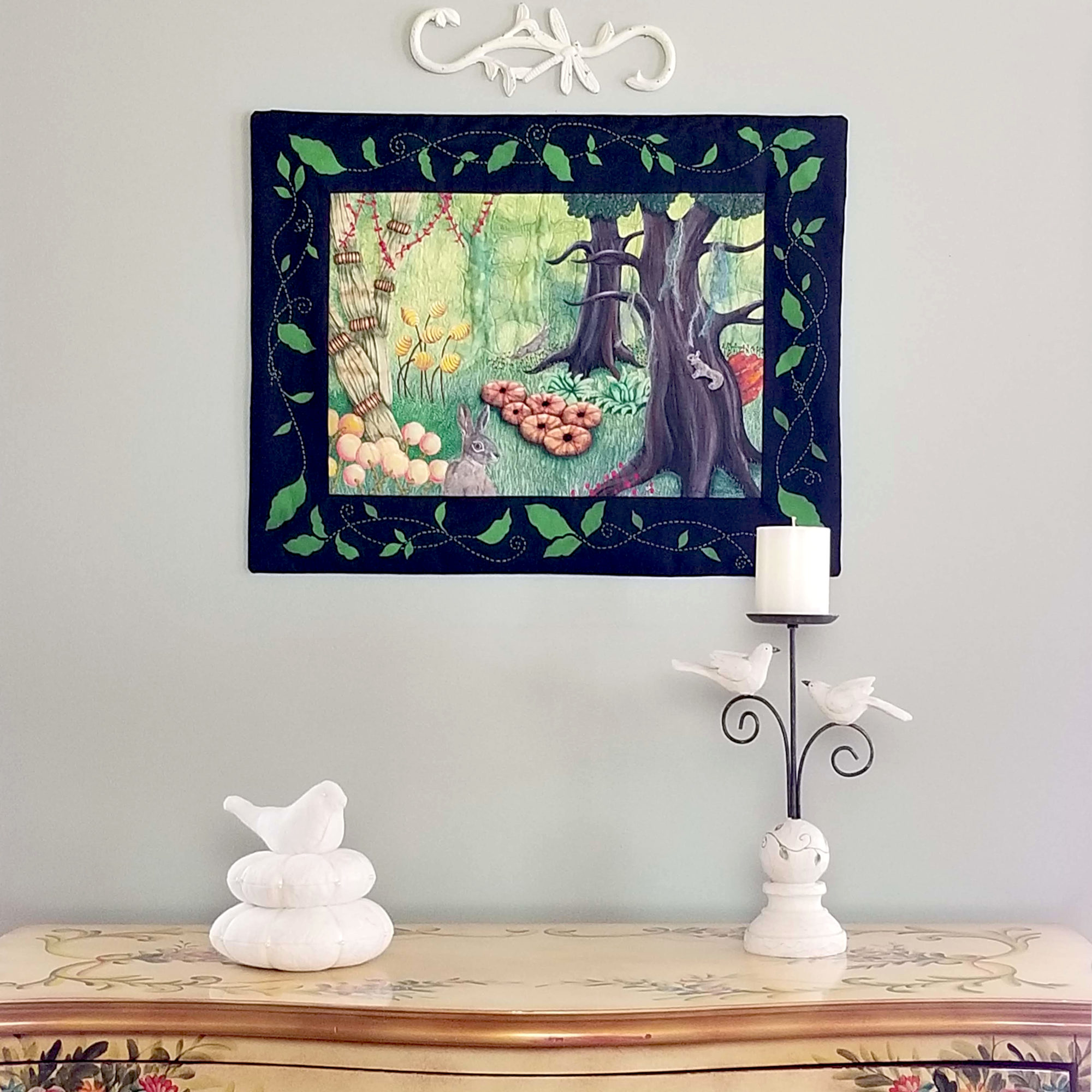 Textile artwork Cache-Cache on a wall above a sideboard where there is a bird on a nest and a candlestick with birds. The artwork shows a scene of 2 hares and a squirrel playing hide and seek in an enchanted forest.