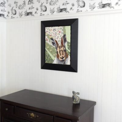 Tableau d'art textile Monsieur Sylvestre encadré dans un cadre noir sur un mur de lambris blanc où il y a au-dessus du papier peint avec des illustrations de lapins noirs sur fond blanc.