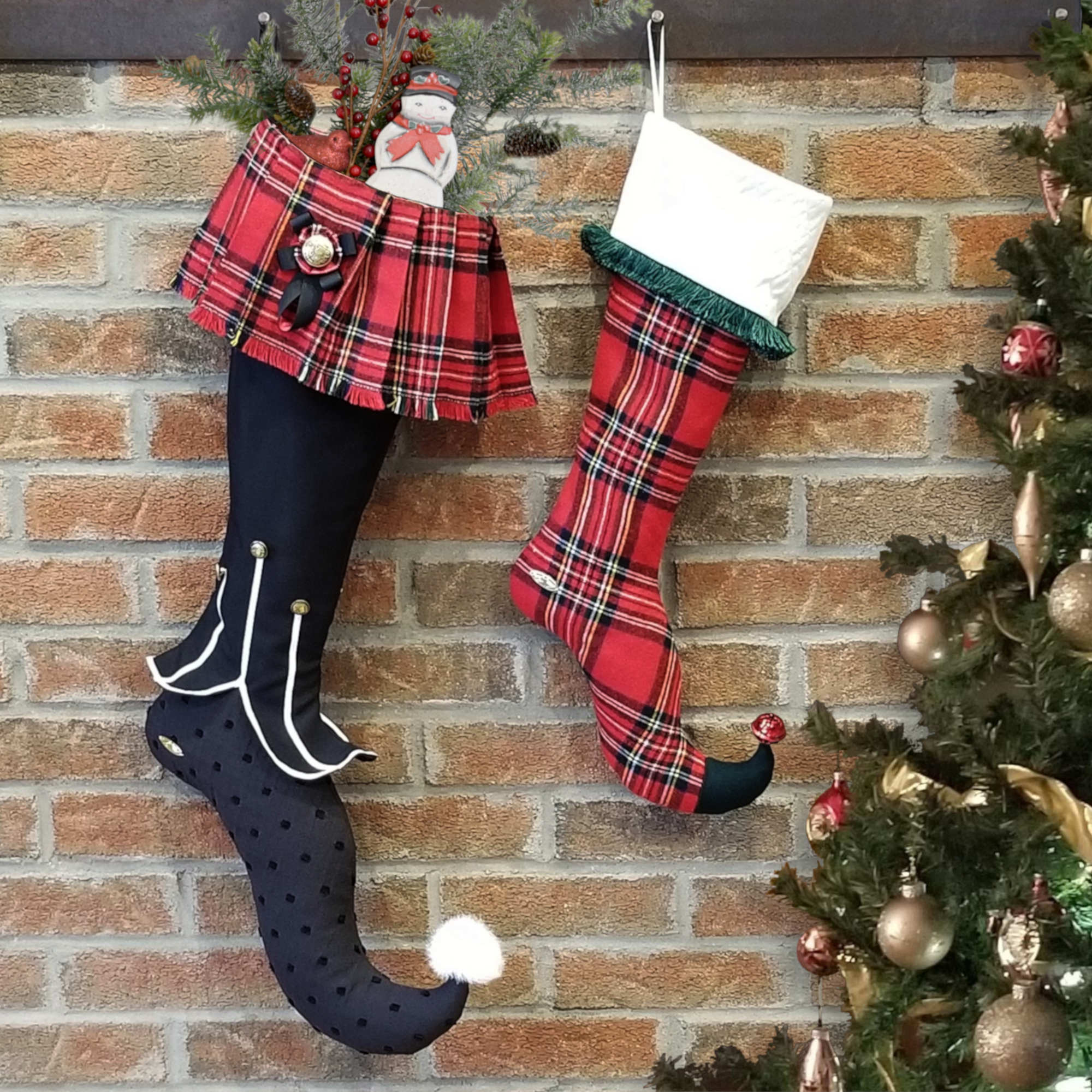 Bas de Noël Scott et Stuart suspendus devant un mur de brique avec un décor de Noël. Les bas sont d'inspiration écossaise. Le bas de gauche en noir et blanc avec un revers en forme de kilt en tartan rouge et le plus petit bas à droite tout en tartan rouge Royal Stewart avec un revers en tissu matelassé blanc cassé et frange vert forêt.