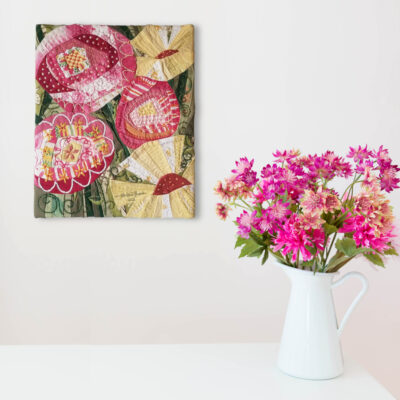 Tableau en tissu représentant des fleurs de couleurs vives roses et jaunes sur un fond vert. Le tout est brodé à la main. Le tableau est sur un mur blanc près d'un bouquet de fleurs roses dans un vase blanc.