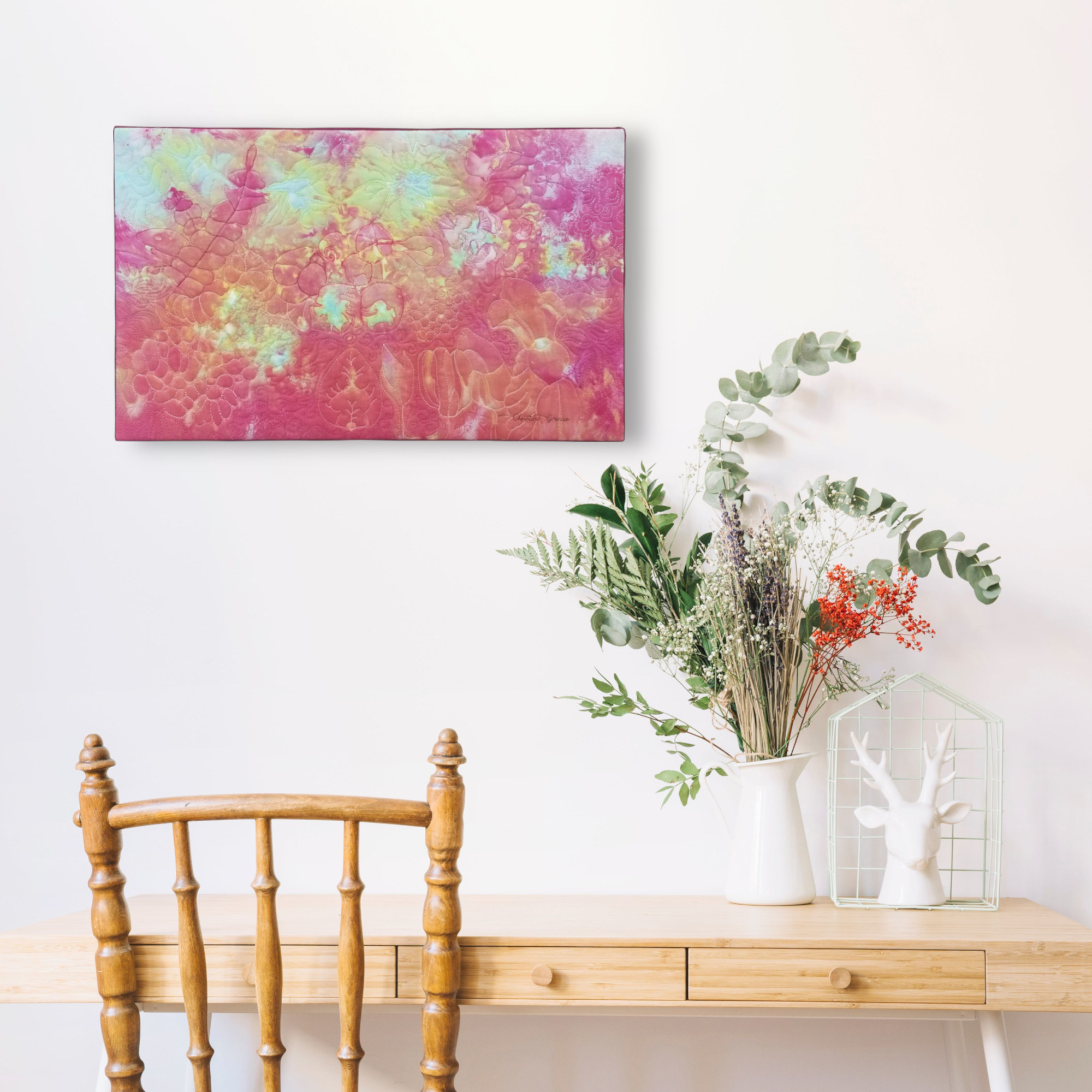 Tableau d'art textile Les jours heureux sur un mur blanc au dessus d'un pupitre avec un bouquet.
